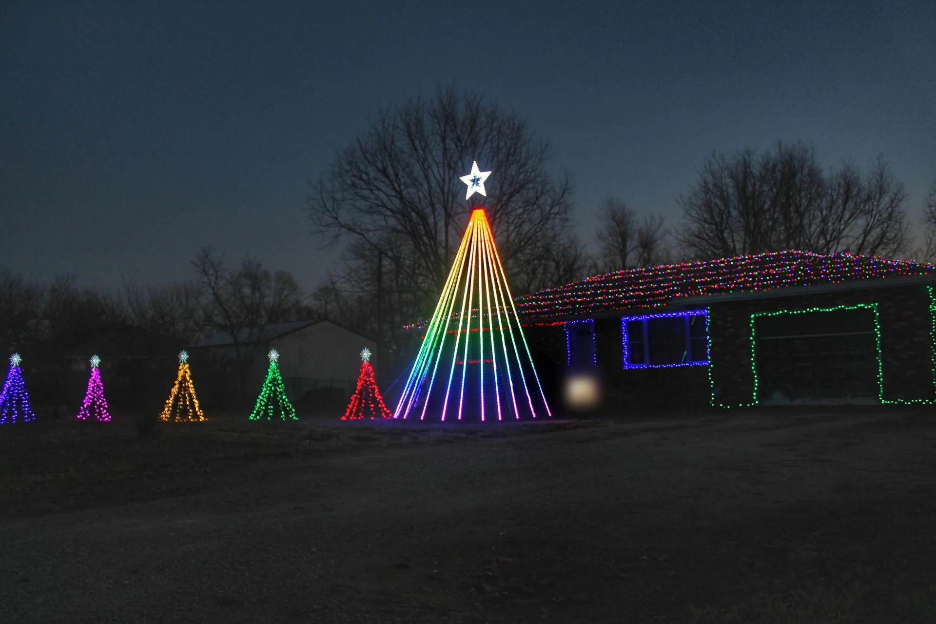 A ranch house at dusk with a rainbow mega tree out front and five small light trees across the yard