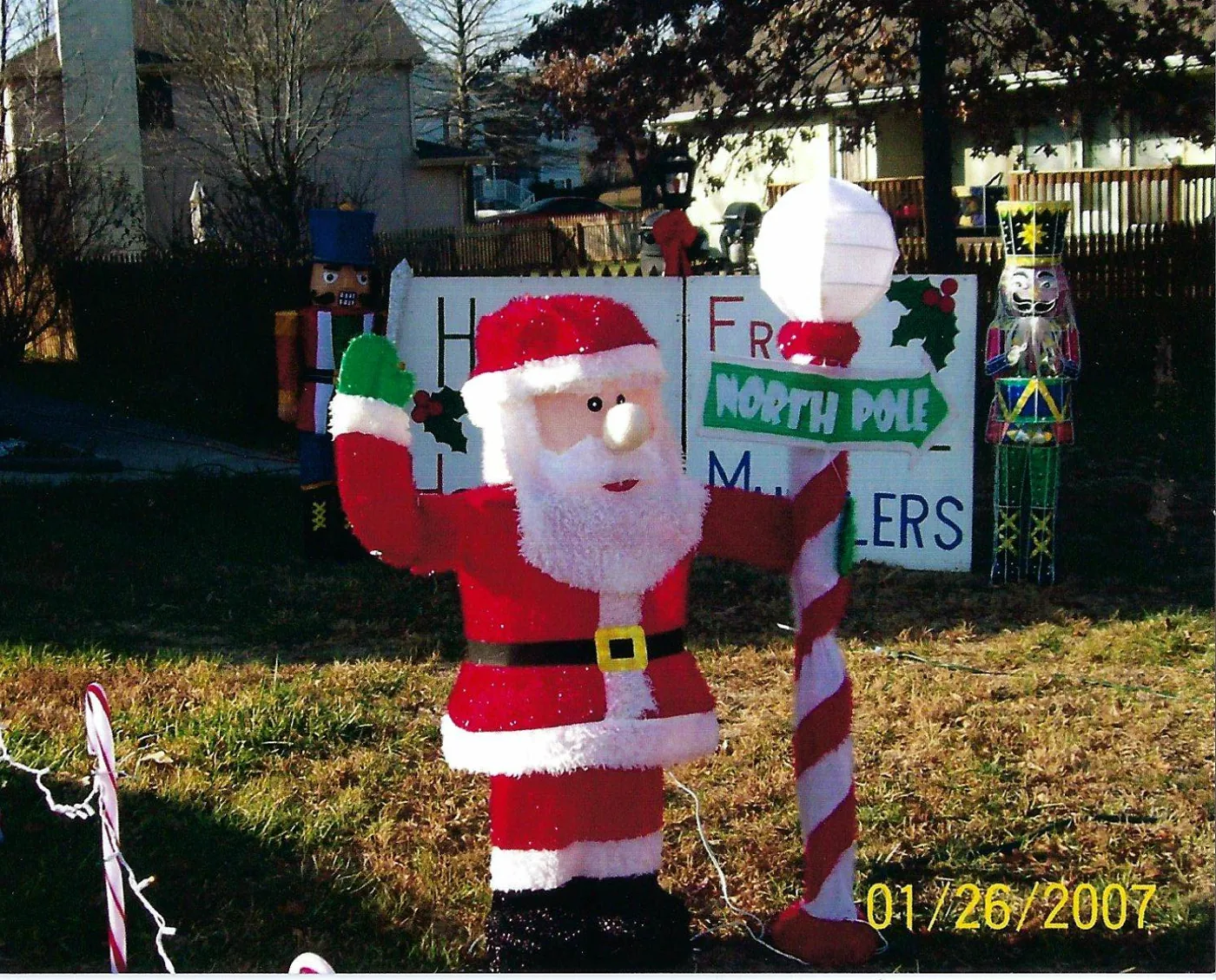A blow mold Santa holding a North Pole sign in front of the Happy Holidays from the Muillers sign